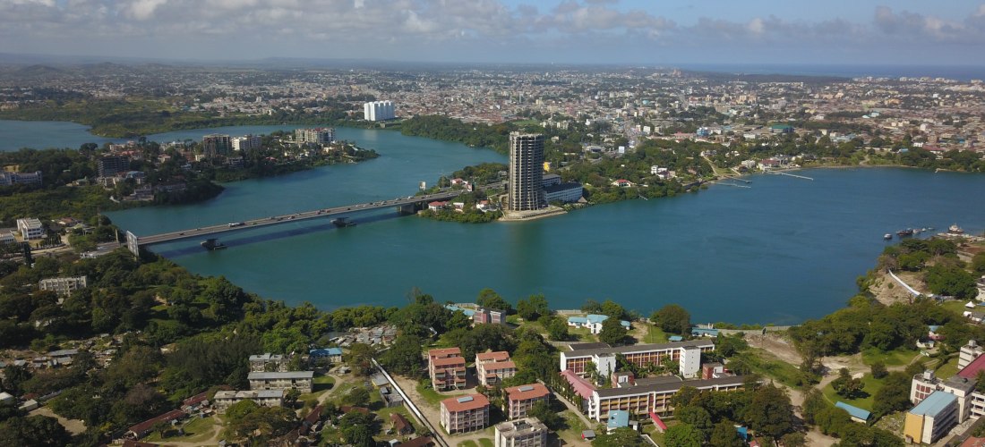 Luftaufnahme von Mombasa, einer Küstenstadt in Kenia. Zu sehen ist eine große Brücke über einem breiten Flussarm, umgeben von Hochhäusern, Wohngebieten und viel Grün. Am Horizont erstreckt sich die Stadt bis zum Meer