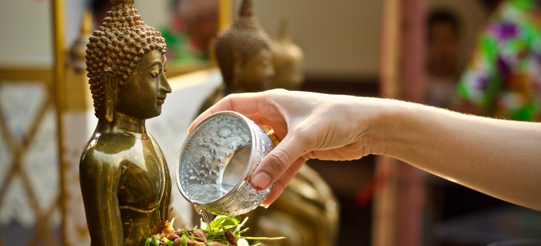 Eine Hand gießt aus einer verzierten silbernen Schale Wasser über eine kleine goldene Buddha-Statue. Vor der Statue liegt ein Blumenopfer aus Jasmin und Rosenknospen.