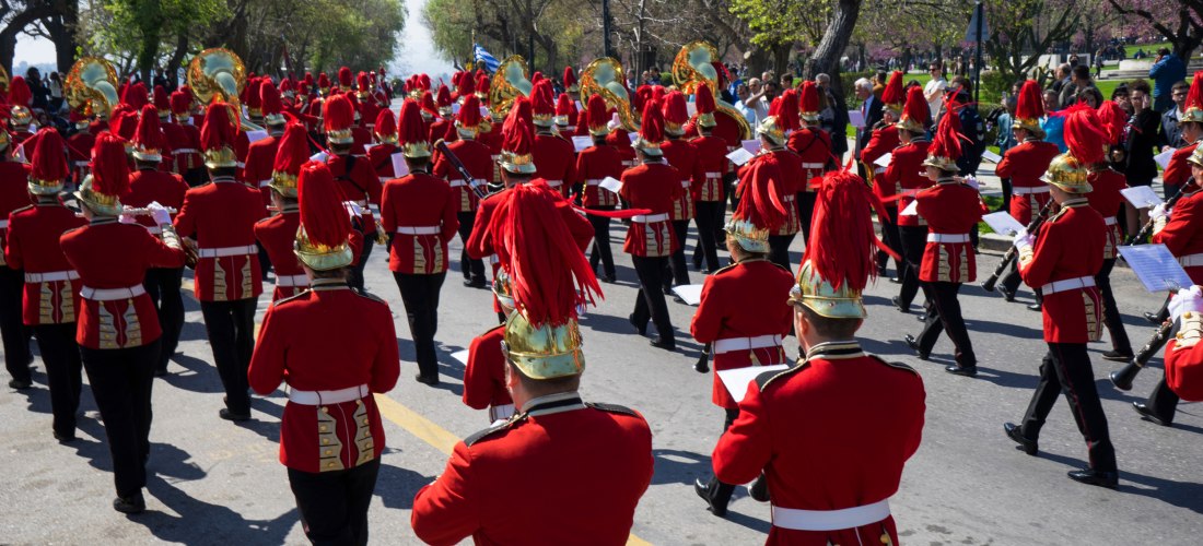 Musik und Tradition auf Korfu: Die Stadtkapelle in markanten roten Uniformen mit goldenen Helmen zieht bei einem festlichen Umzug durch die Straßen. Die Kapelle spielt eine zentrale Rolle bei Feierlichkeiten und verleiht Festen wie Nationalfeiertagen eine