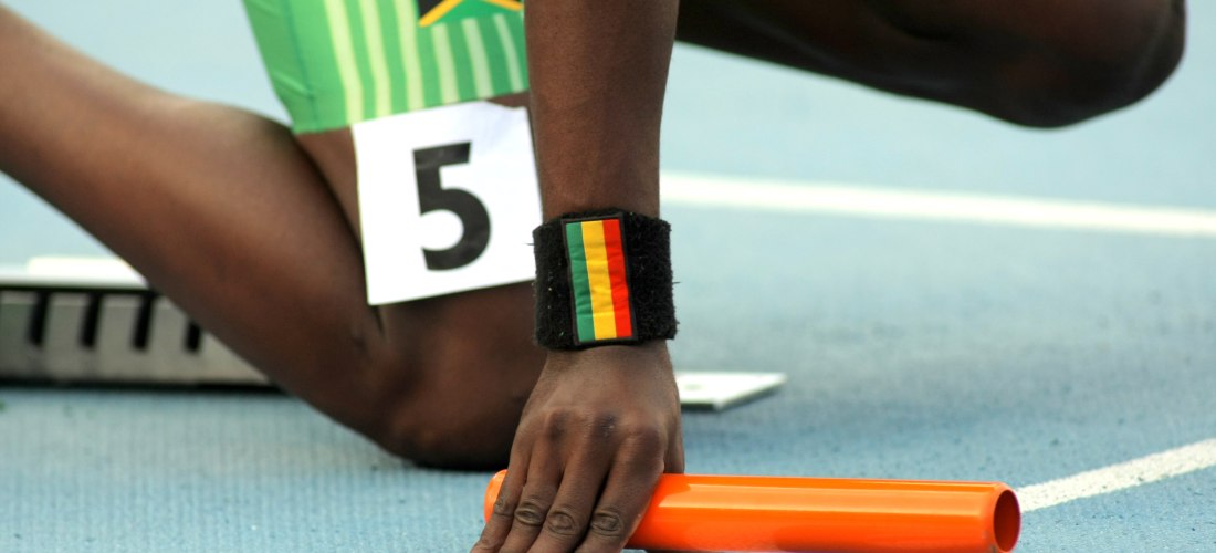 Ein jamaikanischer Leichtathlet in Startposition für ein Staffelrennen auf einer blauen Laufbahn. Der Sportler trägt grüne Laufkleidung mit der Flagge Jamaikas und die Startnummer 5. Am Handgelenk trägt er ein schwarzes Schweißband mit den Farben Rot, Gel