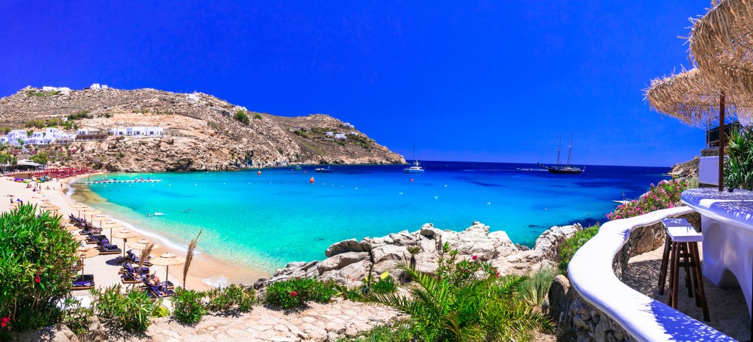 Traumhafter Sandstrand mit türkisblauem Wasser und sanft ansteigenden Hügeln im Hintergrund. Entlang des Strands stehen Liegen und Sonnenschirme. Rechts im Bild eine weiße Barterrasse mit Hockern und einem Strohdach. Auf dem Meer segeln Yachten und ein gr