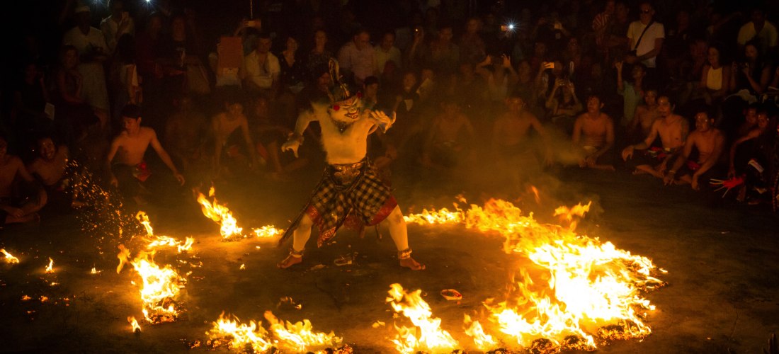 Ein traditioneller balinesischer Tänzer mit weißer Körperbemalung, Maske und kariertem Sarong führt mitten in einem brennenden Kreis einen dramatischen Feuertanz auf. Rundherum sitzen Zuschauer im Dunkeln, viele mit gezückten Handys. Das Bild zeigt eine S