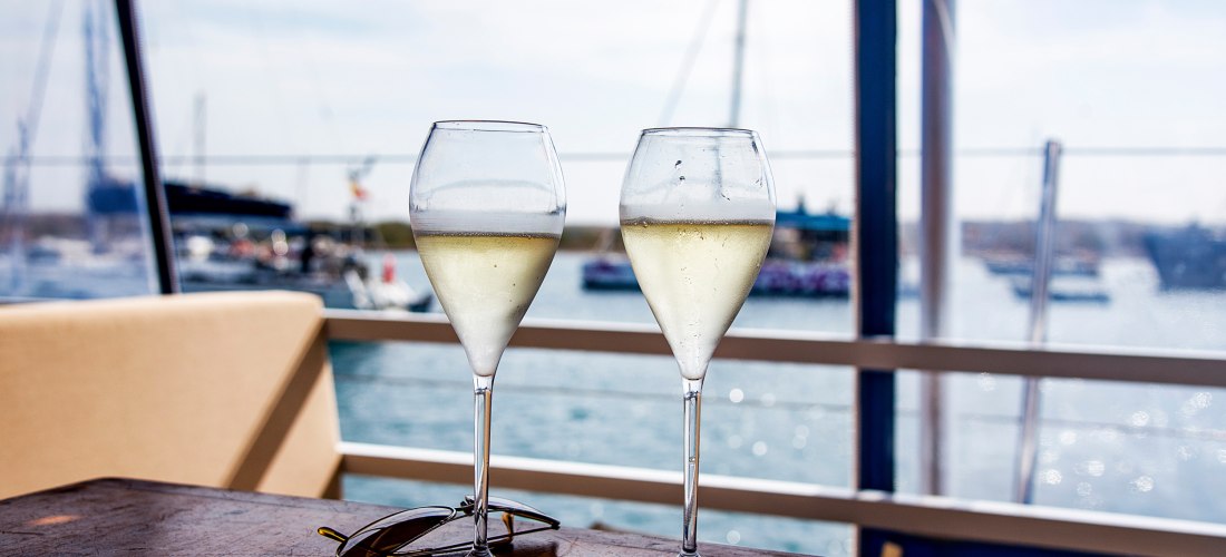 Zwei gefüllte Weingläser mit gekühltem Weißwein stehen auf einem Holztisch in einem Hafenrestaurant. Im Hintergrund verschwimmen Segelboote und Wasser im Sonnenlicht. Eine Sonnenbrille liegt neben den Gläsern auf dem Tisch.