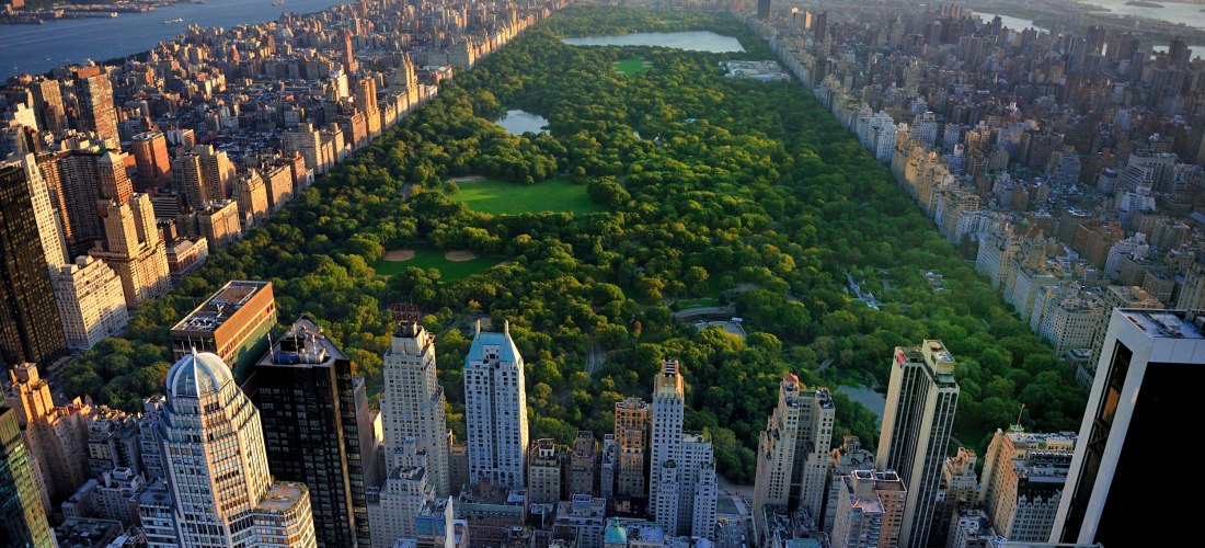 Blick aus großer Höhe auf New York City mit dem Central Park im Zentrum, eingerahmt von Hochhäusern. Links und rechts des Parks erstrecken sich dicht bebaute Stadtviertel. Der Hudson River ist im Hintergrund zu sehen, die Sonne steht tief.