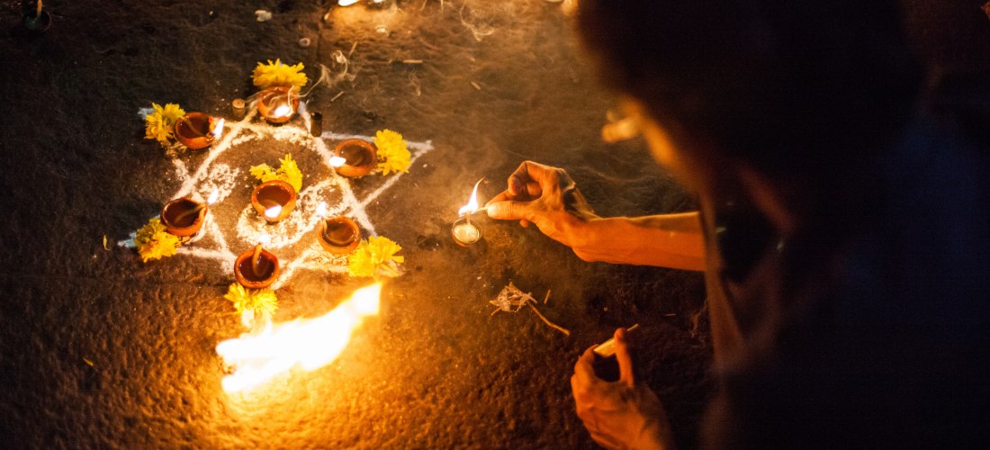tief spirituelle Szene während eines hinduistischen Festes, vermutlich Diwali, dem Lichterfest. Eine Person ist dabei, eine kleine Öllampe (Diya) anzuzünden, umgeben von vielen weiteren brennenden Lichtern. Der Boden ist kunstvoll mit weißen Mustern aus R