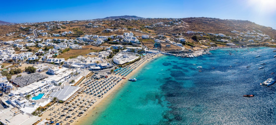 Weitläufige Luftaufnahme einer Küste mit goldgelbem Sandstrand, Reihen von Sonnenschirmen und einer kleinen Stadt mit weißen Häusern. Im türkisfarbenen Meer liegen zahlreiche Boote. Im Hintergrund sanfte Hügel unter klarem, blauem Himmel