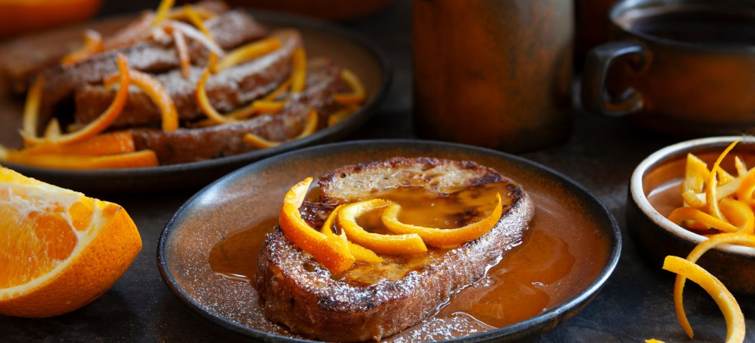Geröstetes Brot in Orangensirup auf einem Teller, garniert mit frischen Orangenzesten. Im Hintergrund weitere Portionen, Orangenscheiben und rustikales Geschirr.