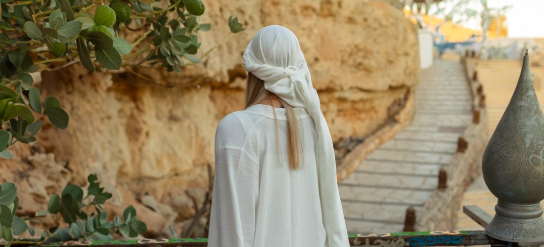 Eine Frau mit langen blonden Haaren steht mit dem Rücken zur Kamera und trägt ein weißes, locker fallendes Gewand und ein Tuch über dem Kopf. Sie blickt auf einen felsigen Weg mit Treppen, der sich entlang einer Steinwand durch eine Wüstenlandschaft schlä