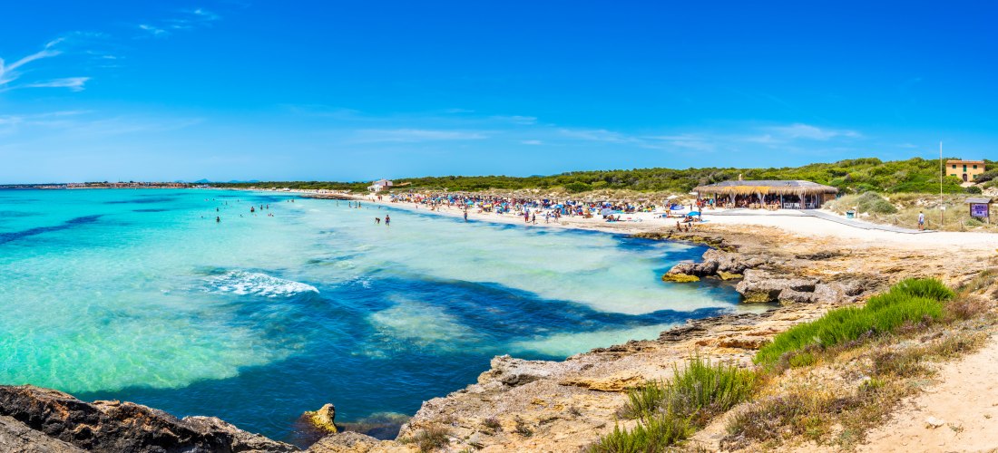 Panorama vom Strand Es Trenc auf Mallorca mit türkisfarbenem Wasser, vielen Badegästen und einem Strandrestaurant im Hintergrund