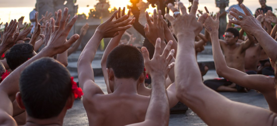 Mehrere balinesische Männer sitzen im Halbkreis und strecken synchron die Arme in die Höhe – Teil einer traditionellen Kecak-Aufführung im Freien. Die Tänzer tragen rot-schwarz-karierte Sarongs und rote Blumen hinter den Ohren. Im Hintergrund versinkt die