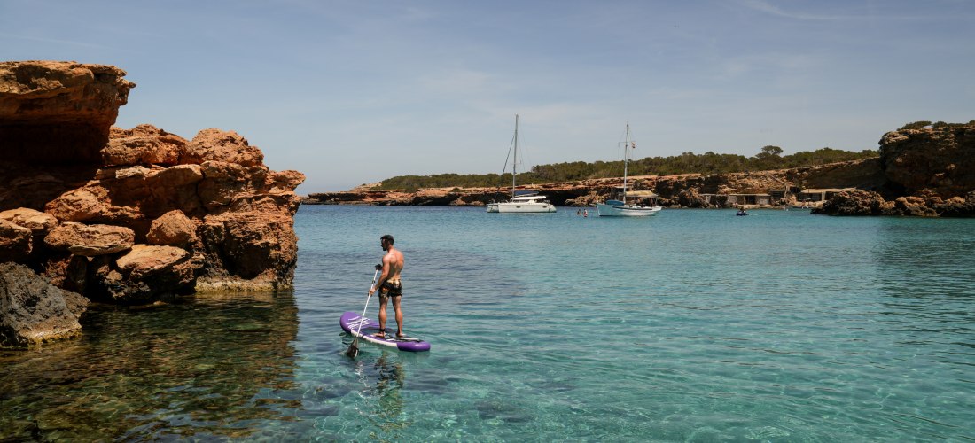 Ein Mann steht auf einem lilafarbenen Stand-Up-Paddleboard auf glasklarem, türkisblauem Wasser. Im Hintergrund liegen Segelboote in einer ruhigen Bucht, umgeben von Felsen und niedriger Vegetation. Der Himmel ist wolkenlos, eine Flugzeugspur zieht sich üb