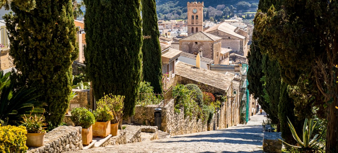 eine Steinstraße unten in der Stadt Pollensa, umgeben von Bäumen und mit Blick auf die Berge