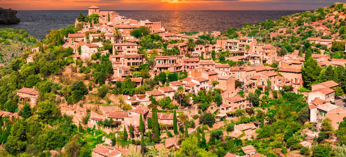 Eine lebendige mediterrane Landschaft mit einer Vielzahl von traditionellen Steinhäusern, die sich an die Hänge eines Hügels schmiegen. Die Häuser sind von vielen Bäumen, Büschen und Gärten umgeben. Im Hintergrund geht die Sonne über dem Meer unter und ta