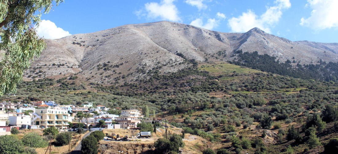 Das Bild zeigt ein ländliches Dorf ( Embona ) am Fuße eines felsigen Gebirges auf einer griechischen Insel, vermutlich Kreta.   Die Häuser sind meist weiß oder hellbeige mit Flachdächern und liegen eingebettet zwischen sanften Hügeln und Olivenhainen.   I