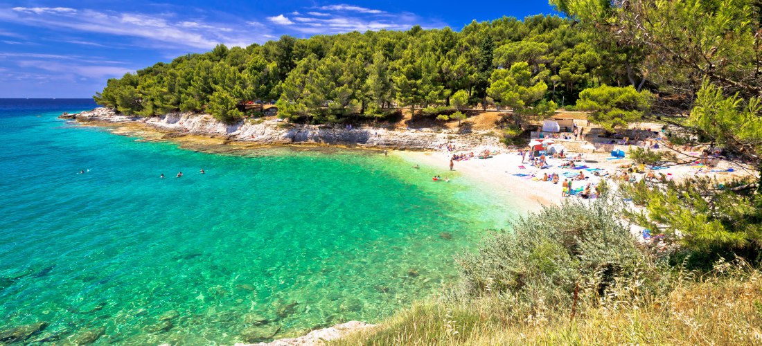 Kroatien - Pula Malerische Badebucht bei Pula in Kroatien mit türkisfarbenem Wasser, hellem Kiesstrand und vielen Badegästen. Im Hintergrund dichter Pinienwald, der bis an das Ufer reicht. Der Himmel ist strahlend blau, einige Menschen schwimmen im Meer oder entspannen a