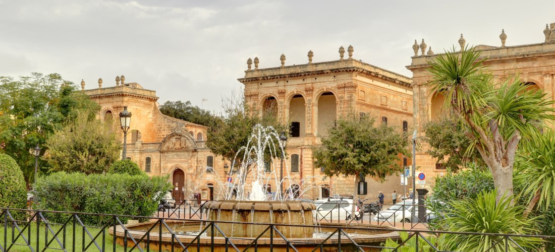 Historische Gebäude aus Sandstein an der Plaça des Born in Ciutadella de Menorca mit Springbrunnen im Vordergrund und Palmen im Park.