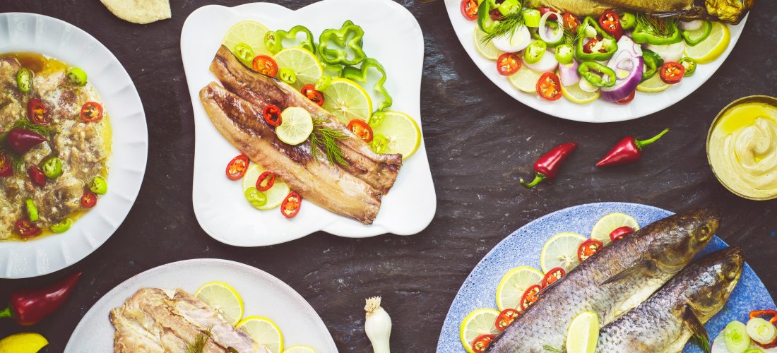 Verschiedene orientalische Fischgerichte auf bunten Tellern, dekoriert mit Zitronen-, Chili- und Zwiebelscheiben sowie frischen Kräutern. Zu sehen sind ganze Fische, Filets und mariniertes Fischfleisch, begleitet von Fladenbrot, eingelegtem Gemüse, Senf u