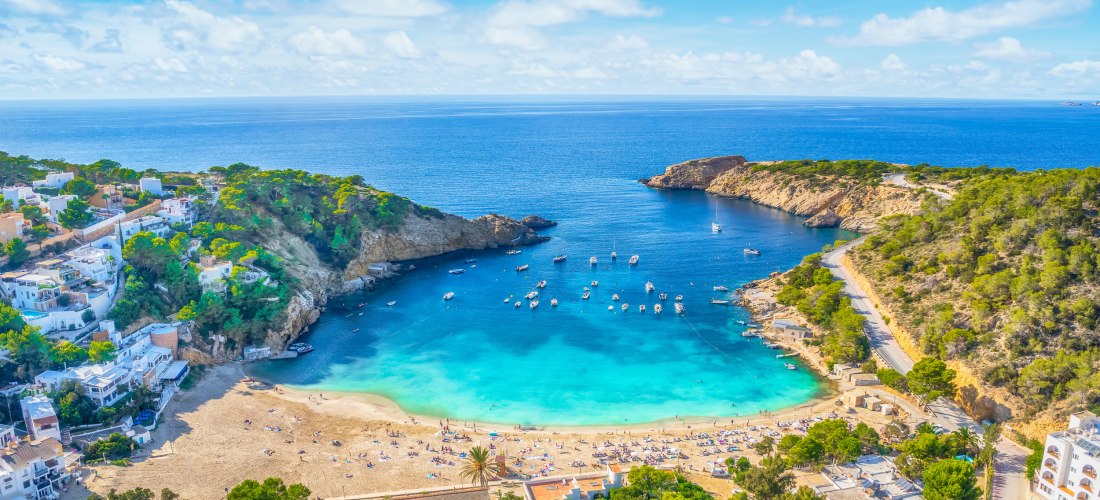Panoramablick auf eine malerische Bucht auf Ibiza. In der Mitte liegt ein sichelförmiger Sandstrand mit vielen Sonnenanbeter:innen. Die Bucht ist von grünen Hügeln mit mediterraner Vegetation und weißen Häusern umgeben. Zahlreiche Boote und Yachten liegen