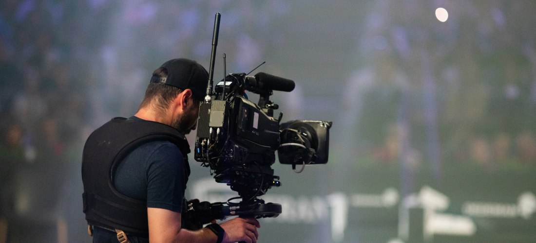 Ein Kameramann in schwarzer Kleidung und Cap trägt ein Steadicam-System mit einer großen Filmkamera bei einer Veranstaltung oder Show mit verschwommenem Publikum im Hintergrund.