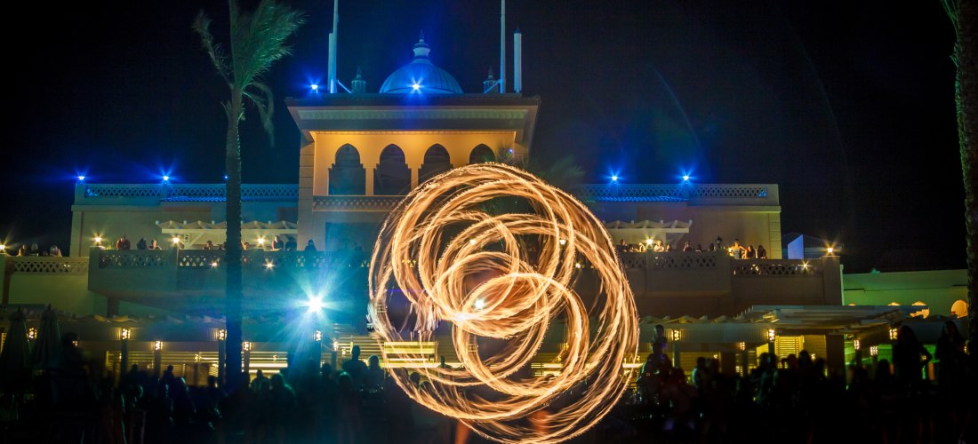 Feuershow bei Nacht: Ein Künstler wirbelt brennende Fackeln, sodass kreisförmige Lichtspuren entstehen. Im Hintergrund ein orientalisches Hotel mit beleuchteten Balkonen und Publikum.