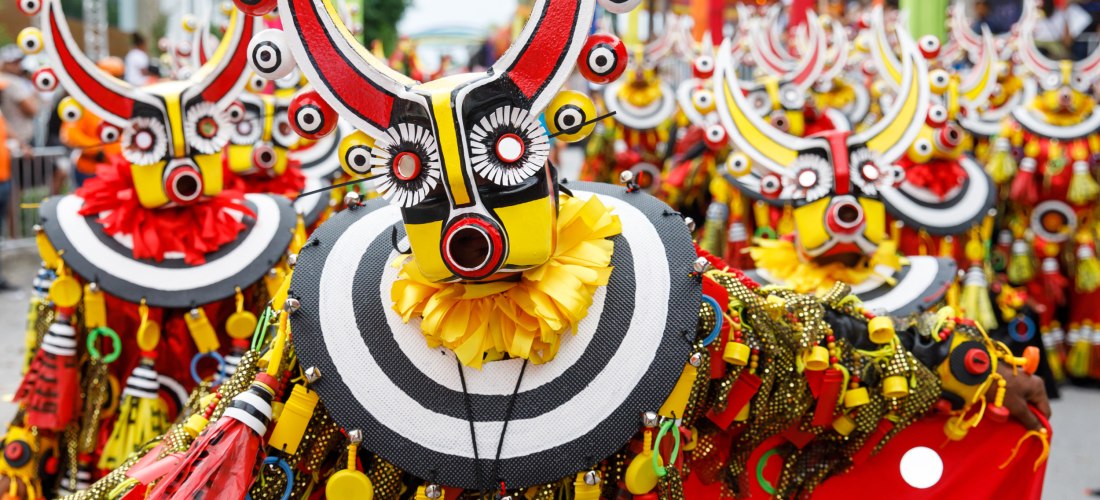 Teilnehmer eines Karnevalsumzugs mit auffälligen Masken in Rot, Gelb und Schwarz, verziert mit Hörnern, Kreisen und Fransen, in einer bunten Straßenparade
