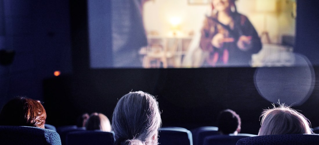 Menschen sitzen in einem Kinosaal und schauen einen Film. Auf der Leinwand ist eine verschwommene Szene mit einer Person in bunter Verkleidung zu sehen. Die Atmosphäre ist ruhig und konzentriert.
