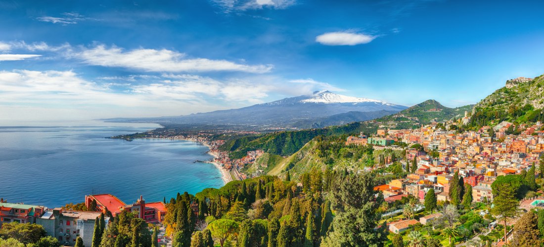 Panoramablick auf die sizilianische Küstenstadt Taormina, die sich terrassenförmig an einen grünen Berghang schmiegt. Rechts im Bild die farbenfrohen Gebäude der Altstadt, mittig der grüne Bergrücken, im Hintergrund der schneebedeckte Vulkan Ätna. Links d