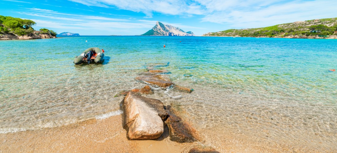 Strand auf Sardinien mit klarem Wasser, Felsen im Sand und Blick auf die Insel Tavolara – beliebtes Reiseziel für Badeurlaub und Naturfreunde