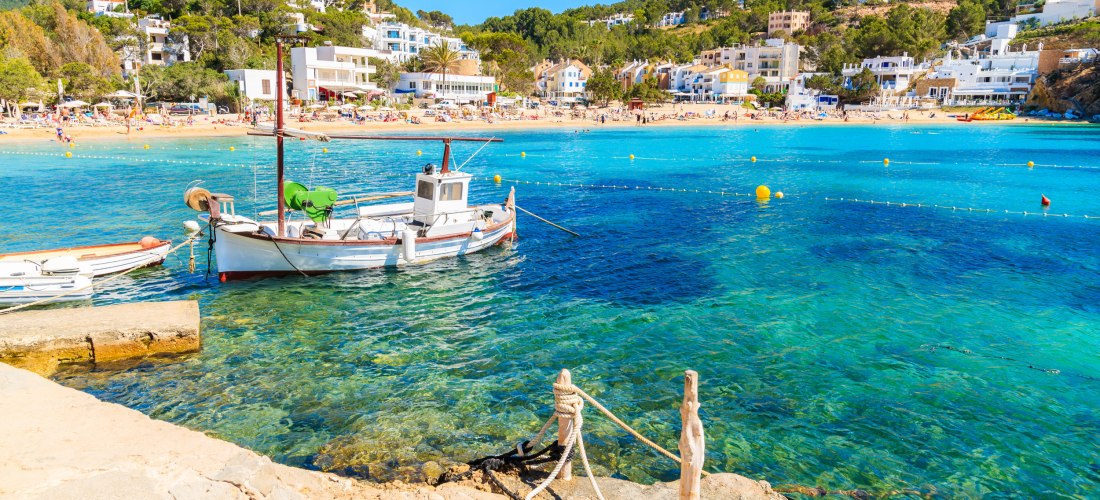 Ein traditionelles weißes Fischerboot mit rotem Mast liegt in einer türkisblauen Bucht auf Ibiza vor Anker. Im Hintergrund erstreckt sich ein belebter Sandstrand mit vielen Menschen, dahinter liegen weiße und pastellfarbene Häuser, eingebettet in grüne Hü