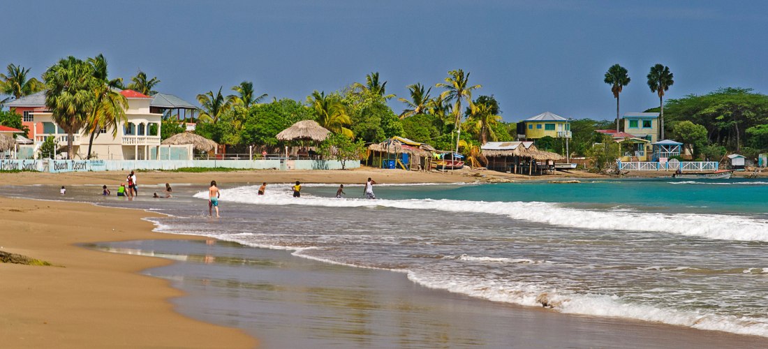 Karibischer Sandstrand mit türkisblauem Meer, Palmen und kleinen Strandhäusern – Badegäste genießen die Sonne und das warme Wasser.