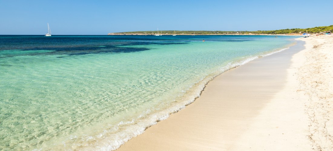 Weitläufiger Sandstrand der Playa de Migjorn auf Formentera mit flach abfallendem, glasklarem Wasser. Im Hintergrund segeln Boote auf dem tiefblauen Meer.