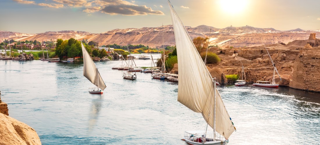 Traditionelle Segelboote mit weißen Segeln gleiten bei Sonnenuntergang über den Nil. Am Ufer sind grüne Palmen, Felsen und antike Steinbauten zu sehen. Im Hintergrund erstreckt sich eine hügelige Wüstenlandschaft unter einem goldfarbenen Himmel