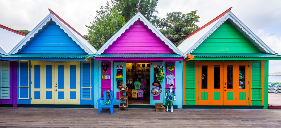 Drei kleine, bunte Holzhäuser im karibischen Stil nebeneinander. Das linke Haus ist blau mit gelben Türen, das mittlere pink-lila mit einem geöffneten Souvenirshop und das rechte grün mit orangefarbenen Türen. Vor dem mittleren Laden stehen bunte Figuren,