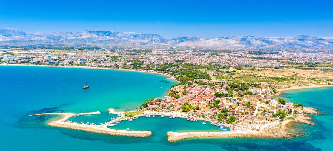 Luftaufnahme einer türkisen Bucht mit einer kleinen Halbinsel, auf der sich eine Altstadt mit roten Dächern und einem kleinen Hafen befindet. Im Hintergrund liegen eine weite Stadtlandschaft und eine Gebirgskette unter klarem, blauem Himmel.