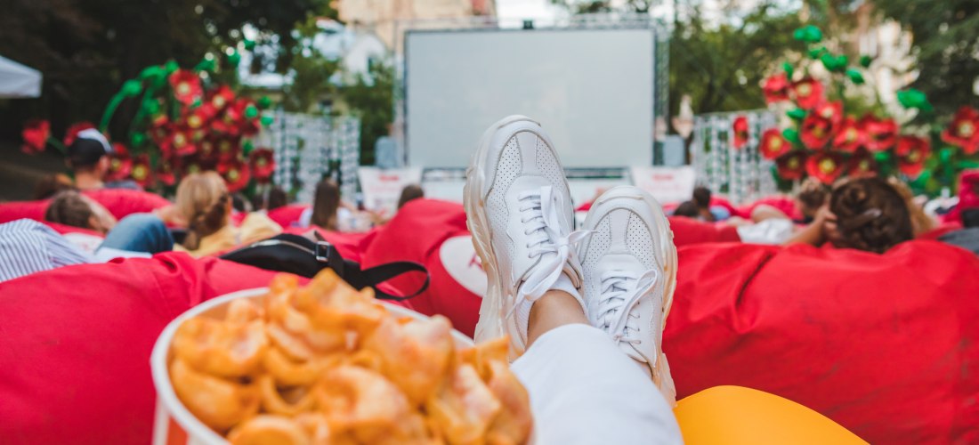 Blick aus der Perspektive einer liegenden Person in einem Open-Air-Kino. Im Vordergrund weiße Sneakers, daneben ein großer Eimer mit Chips. Viele Zuschauer liegen entspannt auf roten Sitzsäcken vor einer großen Leinwand. Die Kulisse ist von Bäumen und dek