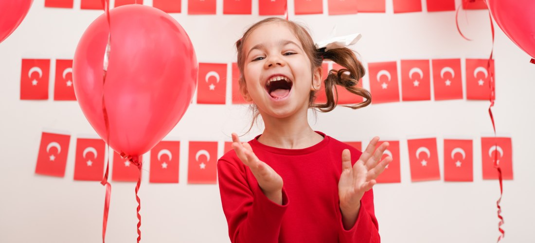 Ein lachendes Mädchen in rotem Pullover steht vor einer Wand mit türkischen Flaggen und roten Luftballons. Sie klatscht in die Hände und wirkt fröhlich und ausgelassen.