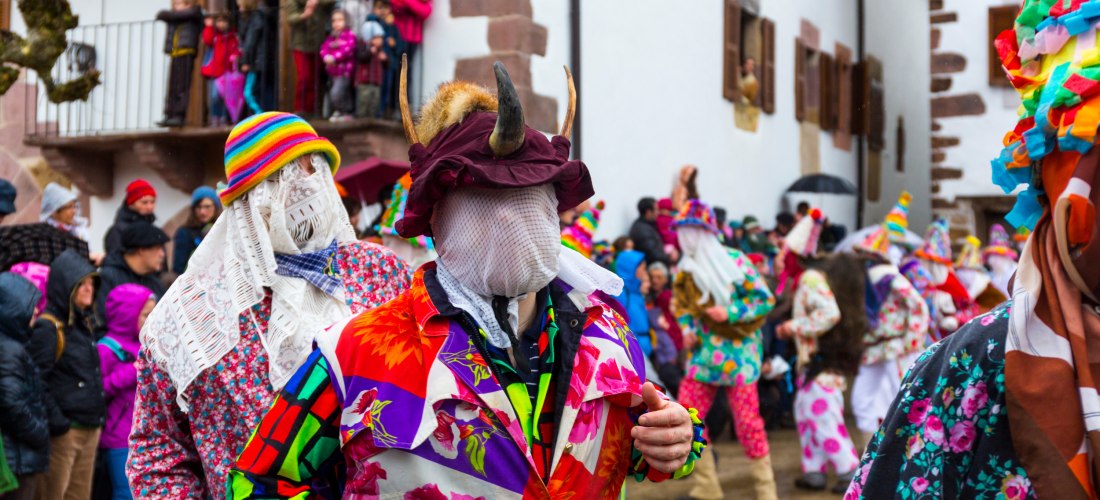 Menschen in auffälligen, bunt gemusterten Kostümen und verschleierten oder mit Hörnern verzierten Masken nehmen an einem Karnevalsumzug teil. Die Straße ist von Zuschauern gesäumt, viele stehen in warmer Kleidung auf Balkonen oder an den Hauswänden. Die V