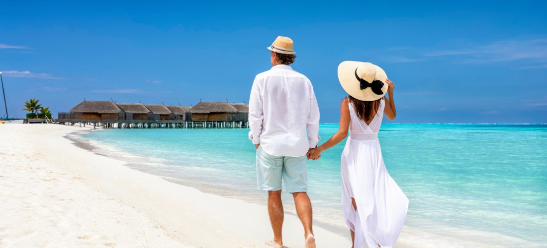 Ein Paar spaziert Hand in Hand am weißen Sandstrand entlang. Im Hintergrund laden luxuriöse Wasservillen              zu einem unvergesslichen Urlaub ein. Perfekt für Zweisamkeit in tropischer Kulisse.