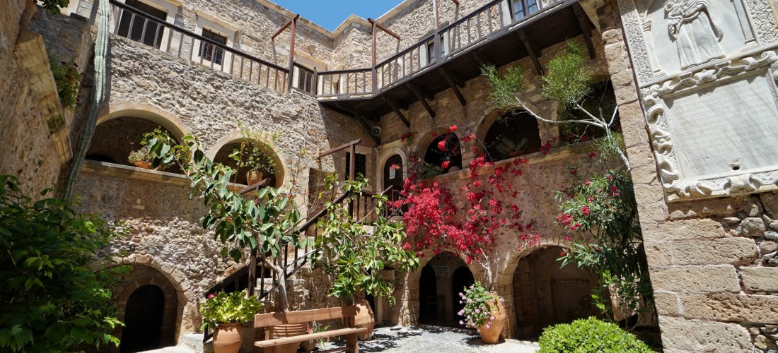 Traditioneller Kloster-Innenhof mit Steinbögen, Holzbalkonen und reich bepflanzten Blumentöpfen. Bougainvillea und Oleander ranken an den Mauern, eine Bank steht im Schatten