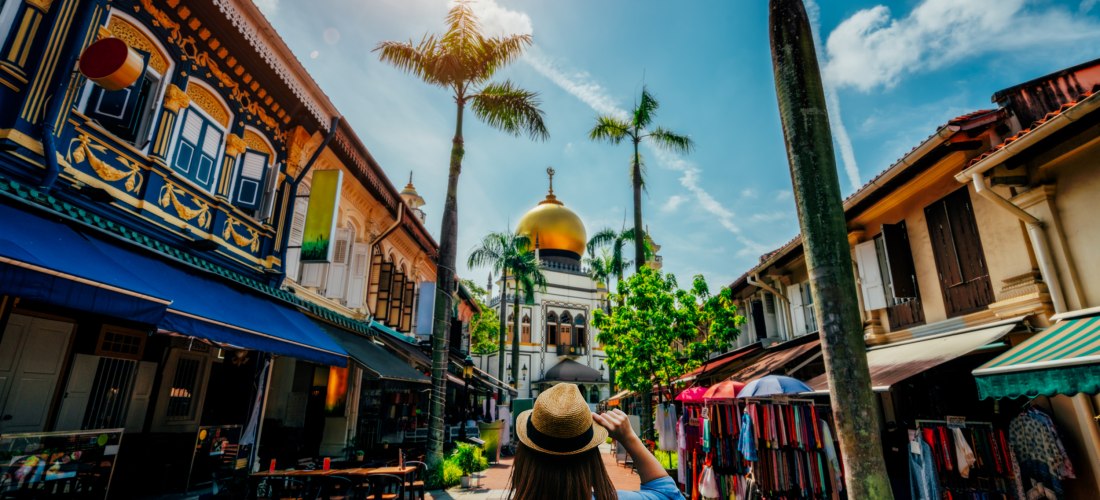 Eine Frau mit Strohhut und Rucksack steht auf einer belebten Fußgängerstraße in Singapur und blickt auf die goldene Kuppel der Sultan-Moschee im Stadtviertel Kampong Glam. Links und rechts säumen farbenfrohe, traditionelle Shophouses mit offenen Fenstern,