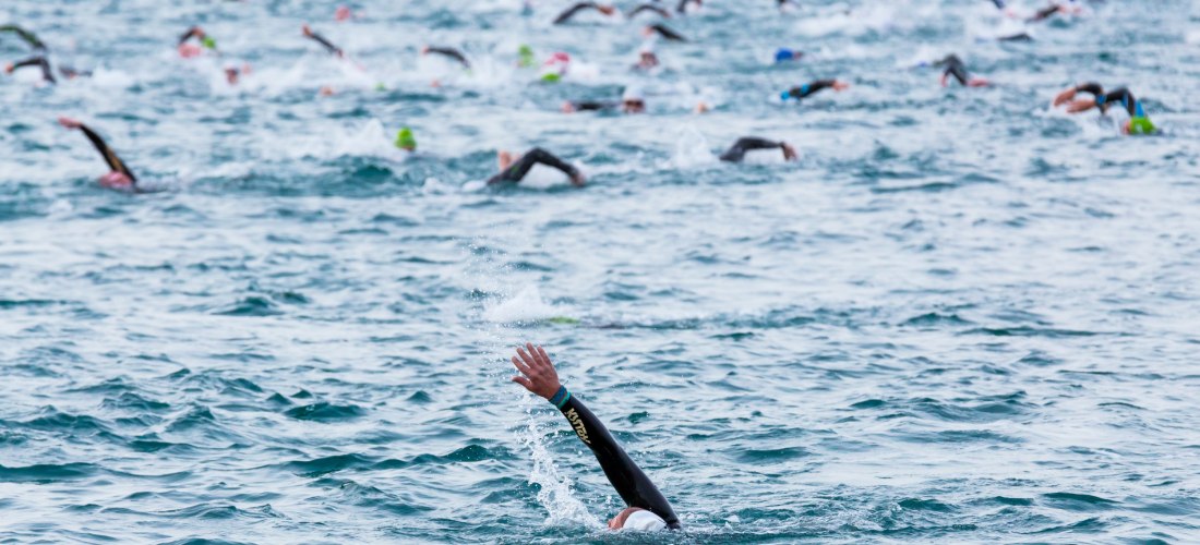Gruppe von Schwimmern in Neoprenanzügen nimmt an einem Freiwasser-Wettbewerb im offenen Meer teil. Eine Person im Vordergrund hebt den Arm beim Kraulschwimmen.