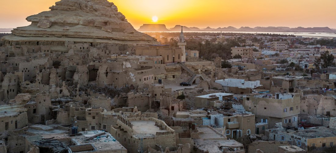 Das Bild zeigt die beeindruckende Lehmbau-Altstadt Schali in der Siwa-Oase, Westägypten, kurz vor Sonnenuntergang. Im Vordergrund erheben sich die verwitterten Überreste der traditionellen Häuser, die aus Salz, Lehm und Palmholz errichtet wurden. Der mark