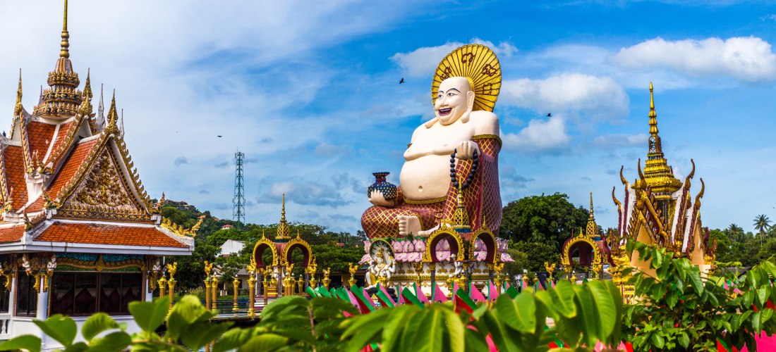 Farbenfrohe Statue des lachenden Buddhas im Wat Plai Laem Tempel auf Koh Samui, Thailand, umgeben von kunstvollen Tempelbauten und tropischer Natur.