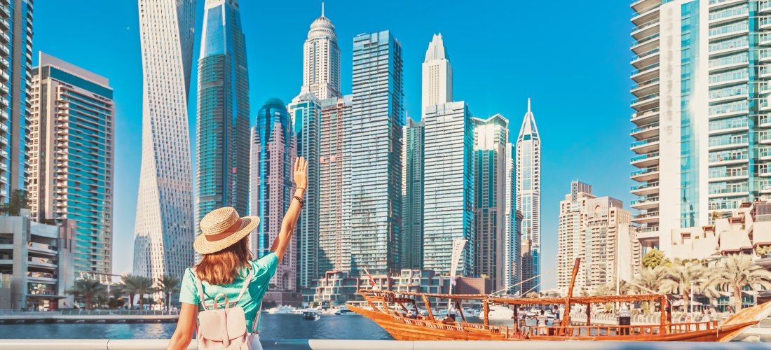 Dubai - Marina Eine Frau mit Strohhut, Rucksack und hellen Sommerkleidern steht an der Promenade der Dubai Marina. Sie blickt auf die Skyline aus modernen Hochhäusern und winkt einem traditionellen Holzboot (Dhau), das über das Wasser fährt. Der Himmel ist klar und stra