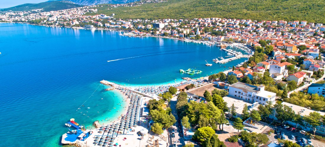 Luftaufnahme einer Küstenstadt mit türkisblauem Wasser, Strand mit Liegestühlen und Wasserrutschen. Im Hintergrund ein grüner Hügel und eine dichte Bebauung mit roten Dächern.