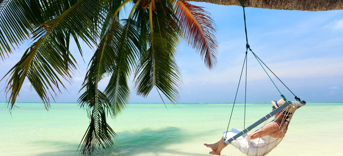 Eine Frau genießt die tropische Ruhe in einer Hängeschaukel unter einer Palme, mit Blick auf das türkisfarbene Meer.              Eine perfekte Szenerie für Entspannung und absolute Erholung am Strand.