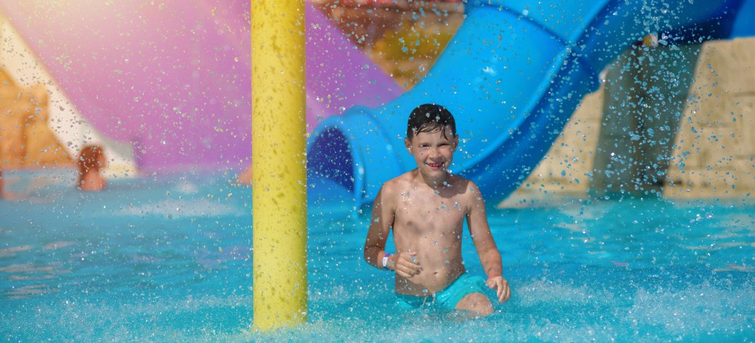 Ein Junge in türkisfarbener Badehose läuft lachend durch das flache Wasser eines Kinder-Wasserparks; Sprühfontänen prasseln herab, im Hintergrund eine blaue Rutsche und bunte Spielelemente.