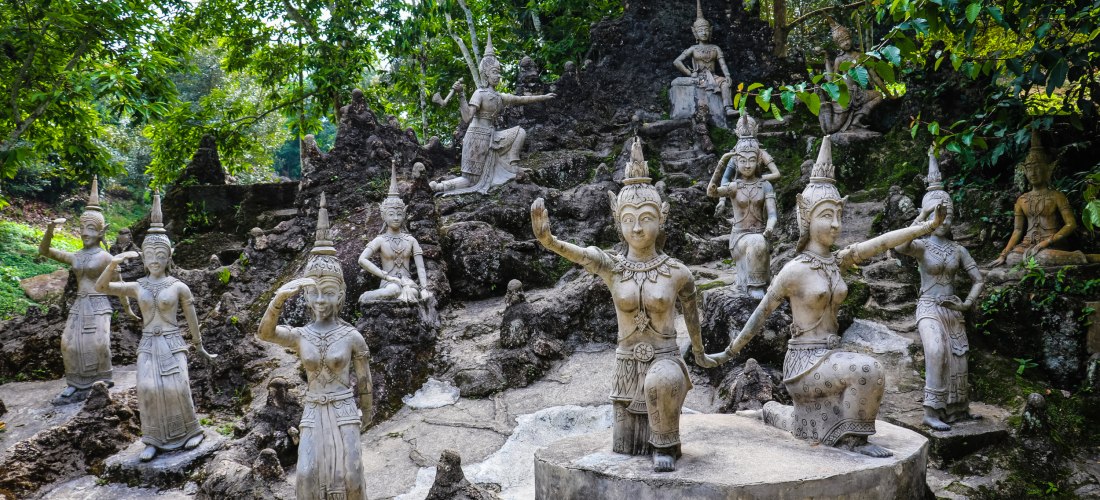 Das Bild zeigt eine Sammlung kunstvoll gearbeiteter thailändischer Steinskulpturen, die in einer felsigen, bewaldeten Umgebung aufgestellt sind. Die Figuren stellen traditionelle Tänzerinnen und mythologische Wesen dar, jede in einer anderen Haltung – ste