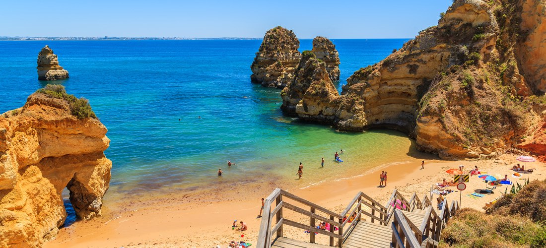 Holztreppe führt hinunter zum idyllischen Sandstrand Praia do Camilo an der Algarve in Portugal. Menschen liegen am goldenen Strand oder baden im türkisfarbenen Meer, das von beeindruckenden Felsformationen umgeben ist. Der Himmel ist klar und strahlend b