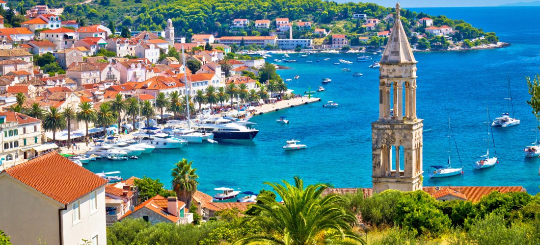 Blick auf den malerischen Hafen von Hvar-Stadt auf der kroatischen Insel Hvar. Entlang der Uferpromenade säumen Palmen die Küste, davor liegen zahlreiche Yachten und Boote im türkisblauen Wasser. Im Vordergrund ragt der historische Glockenturm einer Kirch